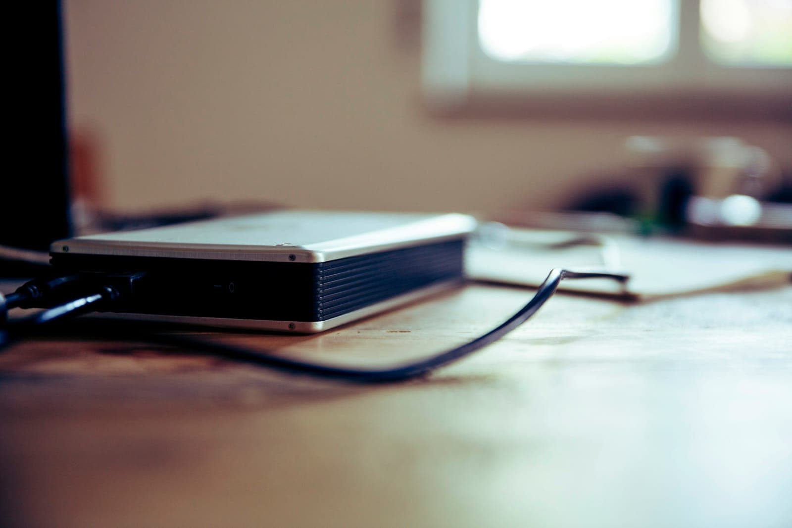 An external hard drive with a silver case sits on a wooden desk, its cables plugged in at one end, with a window and papers blurred behind it.