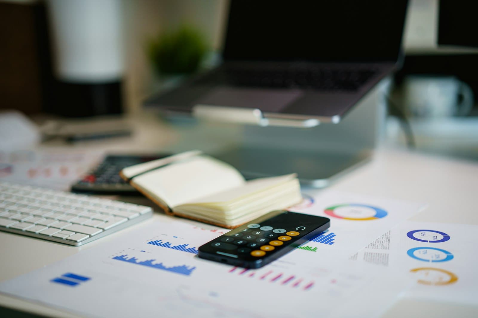 يظهر مكتب عليه أوراق مطبوعة فيها رسوم بيانية، وهاتف يعرض آلة حاسبة، ودفتر مفتوح، ولوحة مفاتيح، ولابتوب على حامل خلفها.