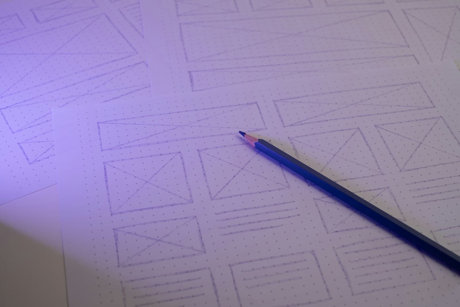Hand-drawn page layouts on dot grid paper, with boxes for images and lines for text, and a blue pencil resting on them.