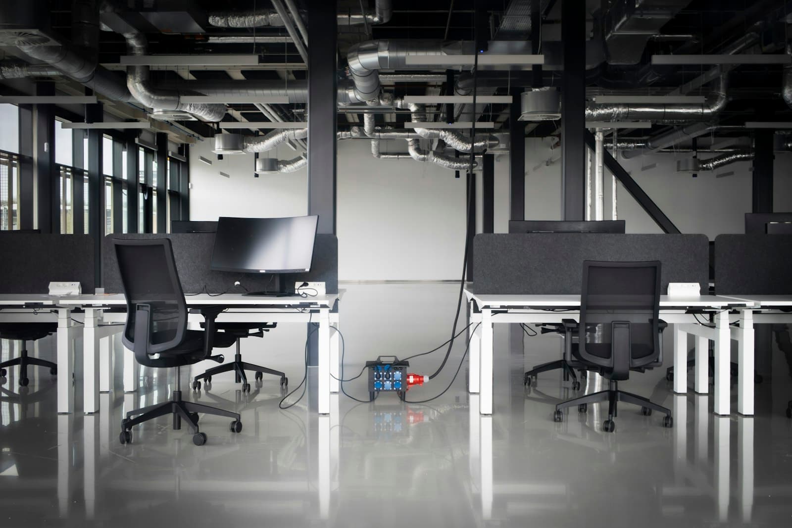An open-plan office still being fitted out: white desks with black chairs and a monitor, an exposed ceiling with ducts, and a temporary power box on the floor.