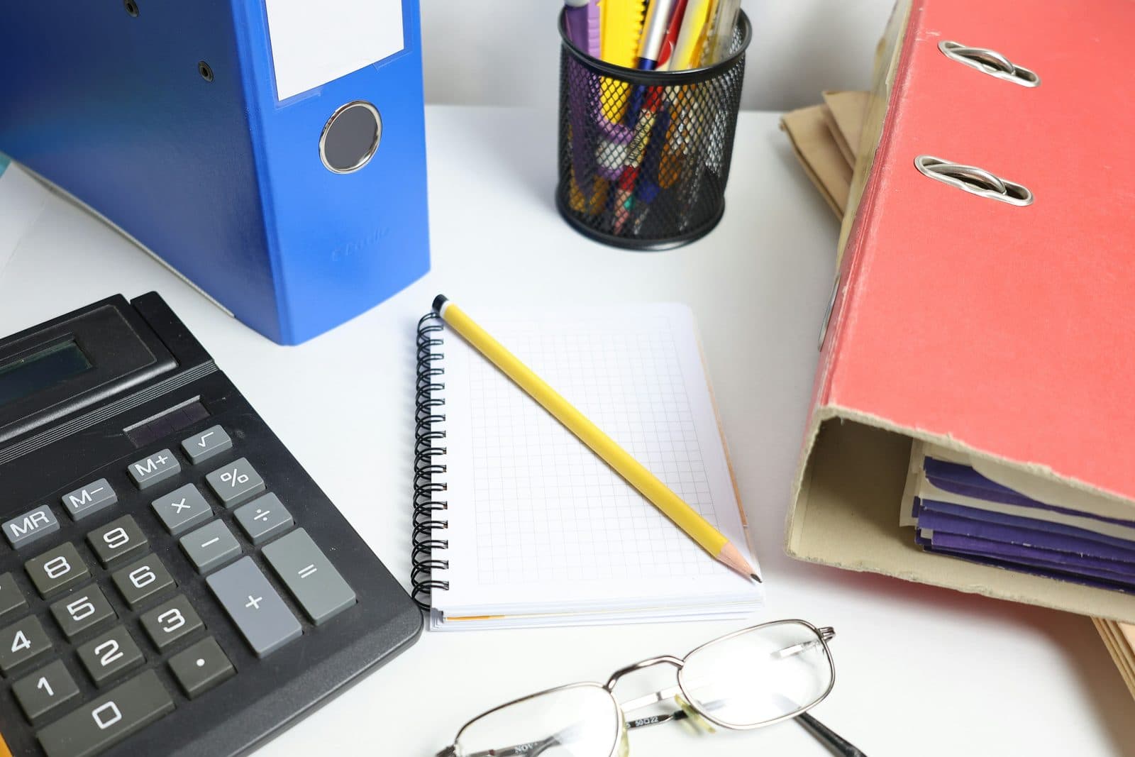 A calculator, ring binders and a squared notebook with a pencil on a white office desk.