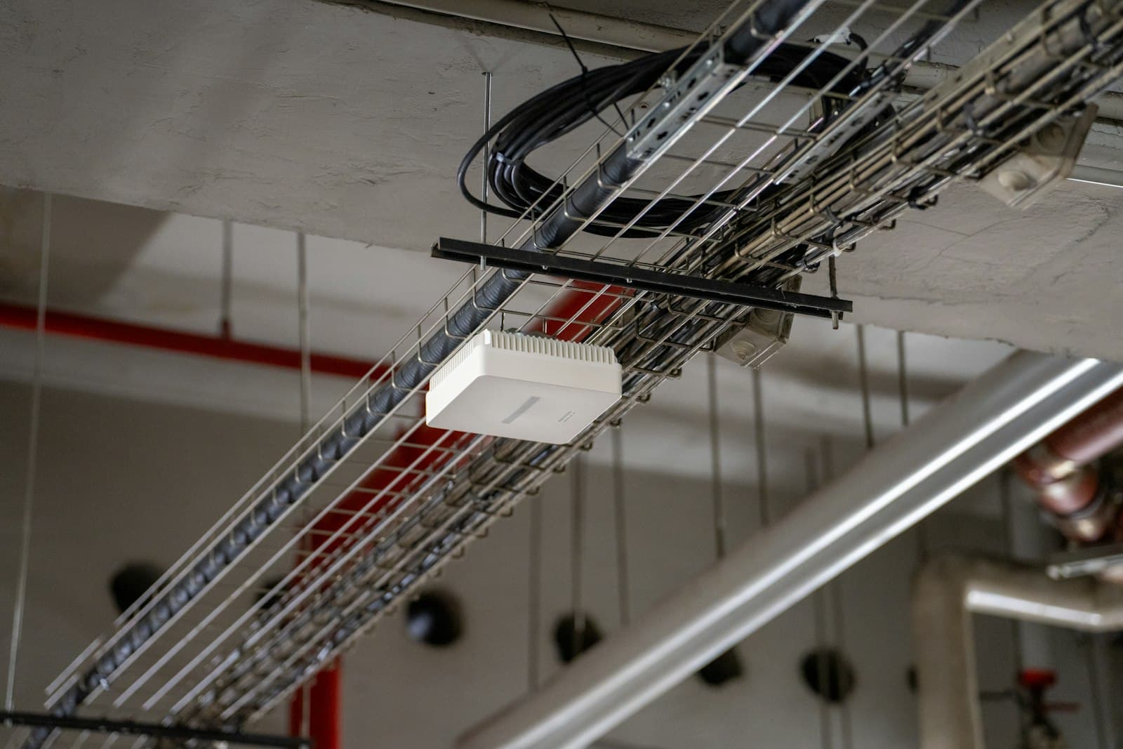 A white Wi-Fi access point fixed to a cable tray under an open ceiling, with network cables running along the tray.