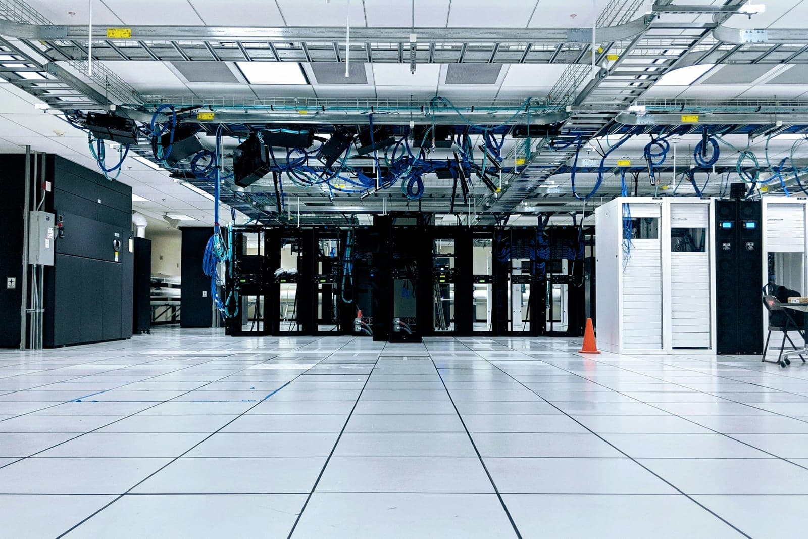 Black racks stand in a row in a bright server room, under ceiling cable trays full of blue network cables, with cooling units at the sides and a white tiled floor.