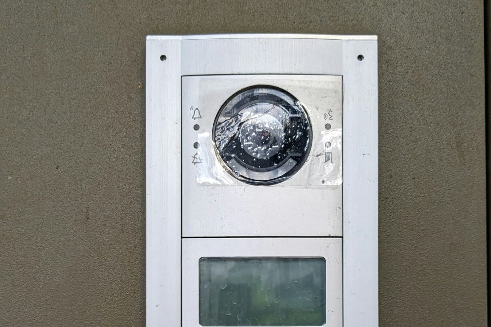 An outdoor video intercom station on a wall, with a camera lens behind a protective film, call and door icons beside it, and a small display below.